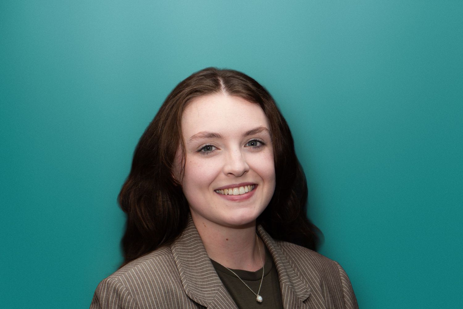 A smiling young woman with brown wavy hair poses against a teal background in a professional headshot.