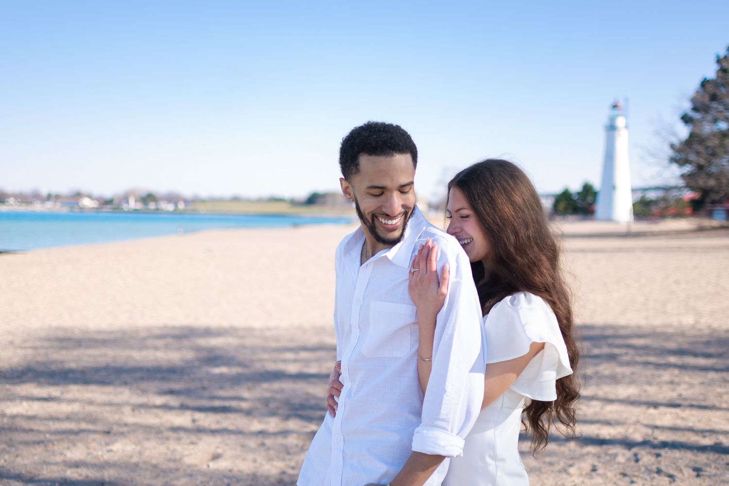 A happy couple in white outfits laughing together on a sandy beach with a lighthouse in the background.