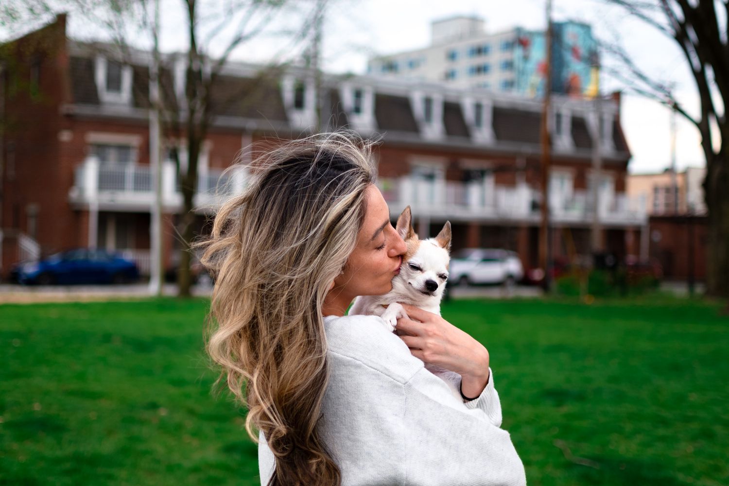 Woman in white sweater kissing a tiny Chihuahua puppy outdoors in a green park with brick buildings in background.