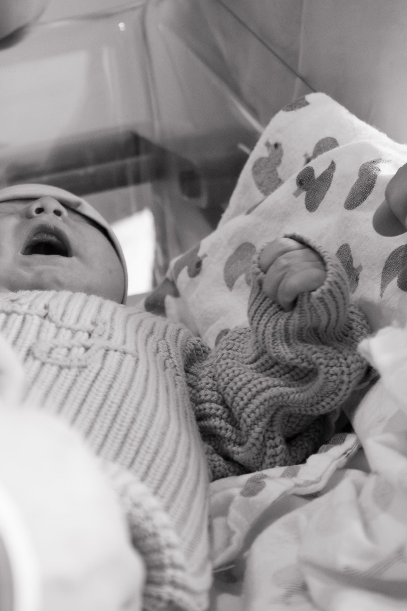 Black and white photo of swaddled newborn baby in knit outfit holding a crocheted toy.