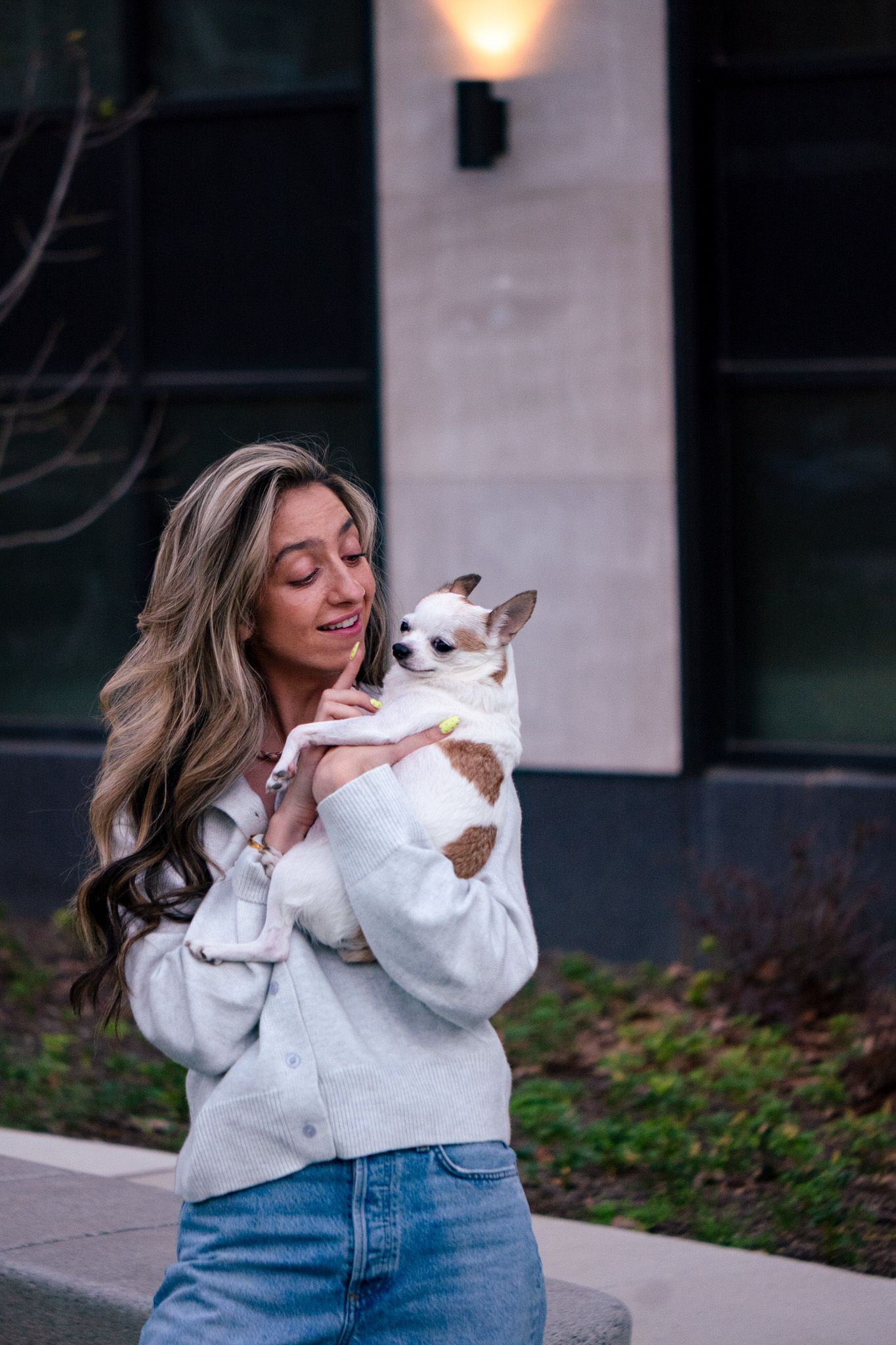 Young woman with long blonde hair holding a small Chihuahua dog while standing outside a modern building.