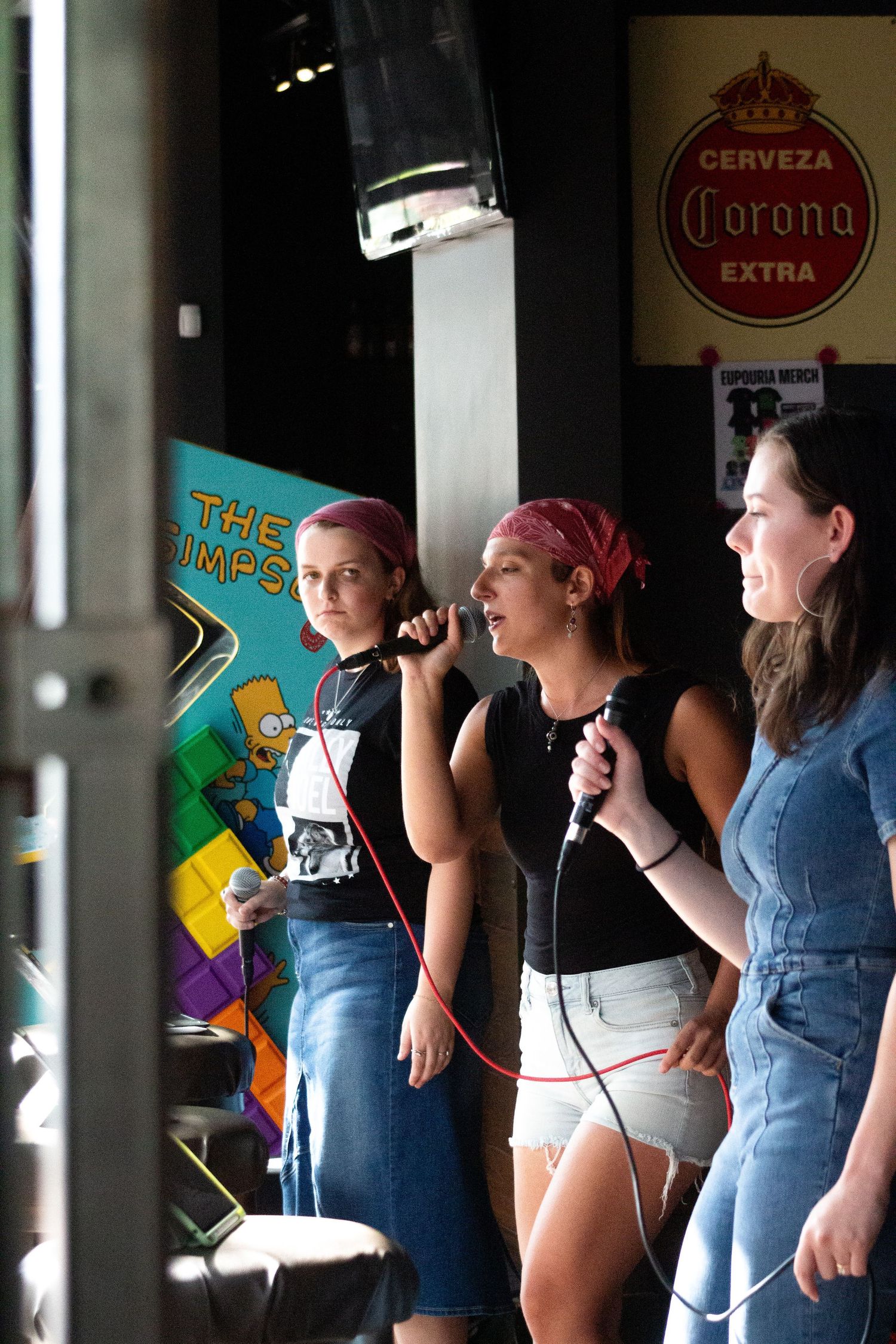 Three female vocalists performing with microphones at live music event near Corona sign.