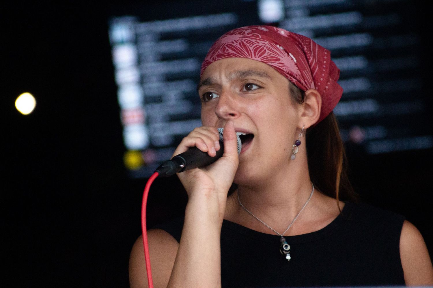 Young woman with red bandana passionately singing into microphone at karaoke night event.