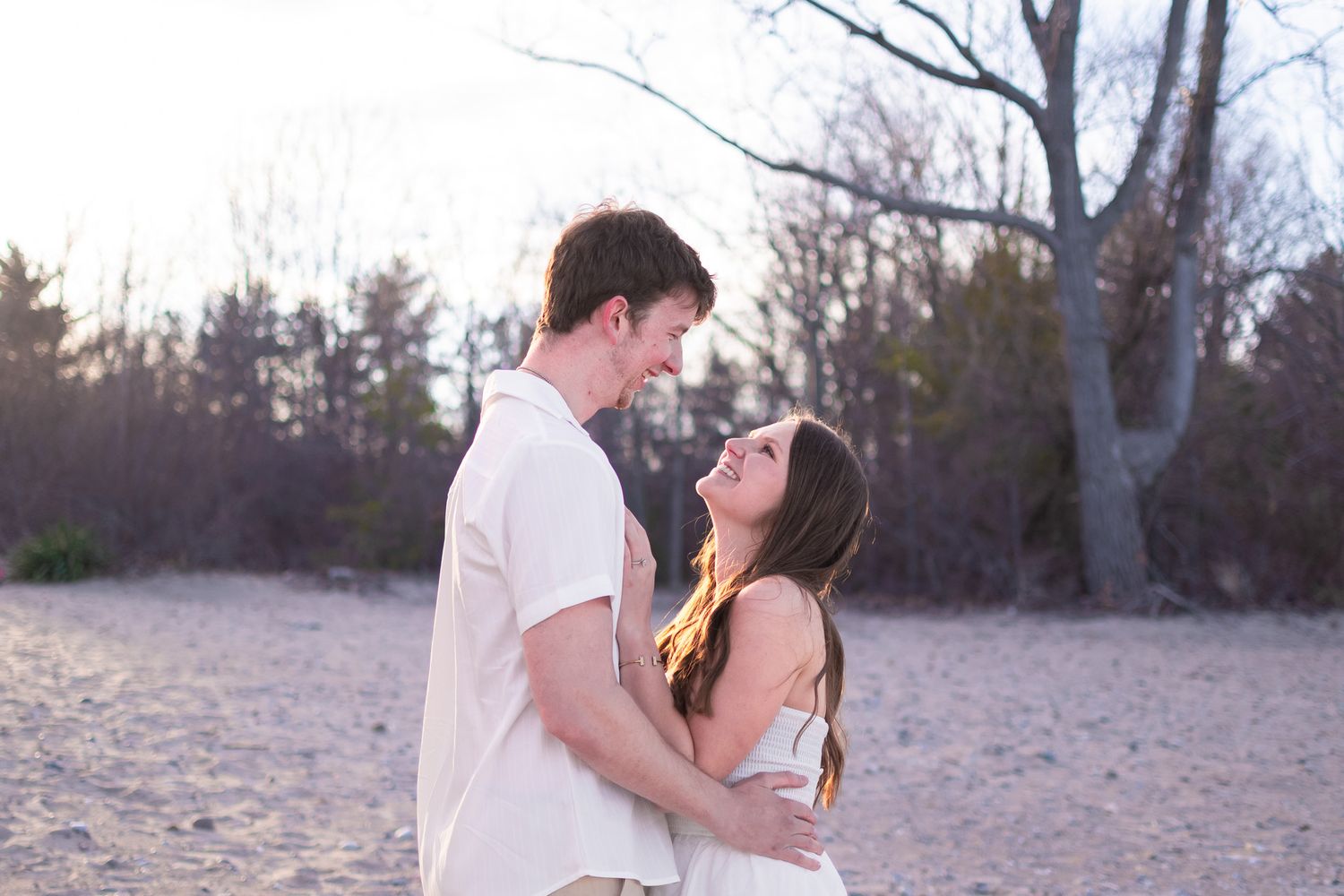 A smiling couple in white outfits embracing on a sandy beach surrounded by bare winter trees at golden hour.