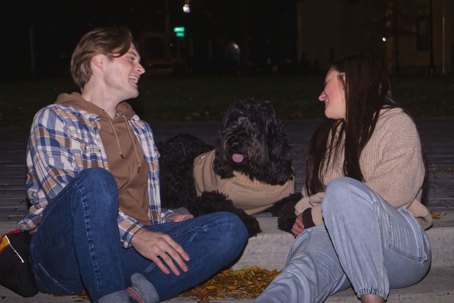 Two young women sitting outdoors at night with a large black dog between them.