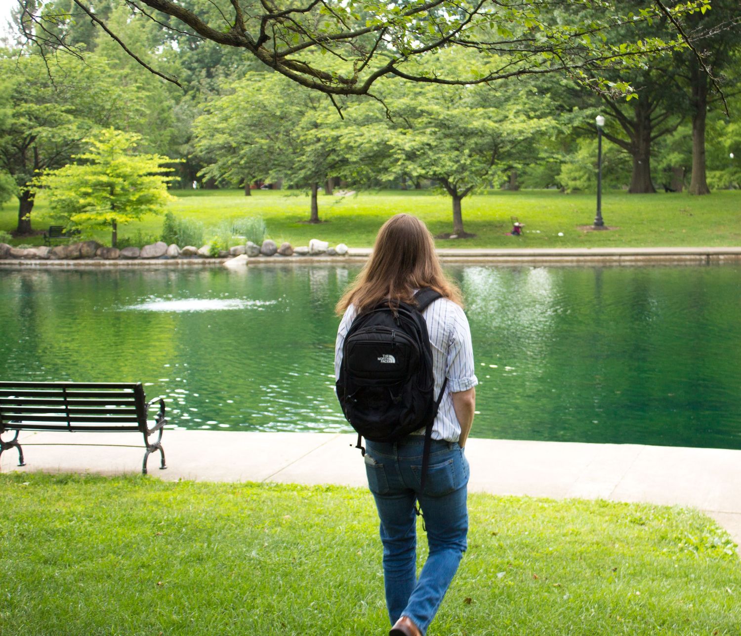 Woman with North Face backpack standing by a serene green park pond under trees.