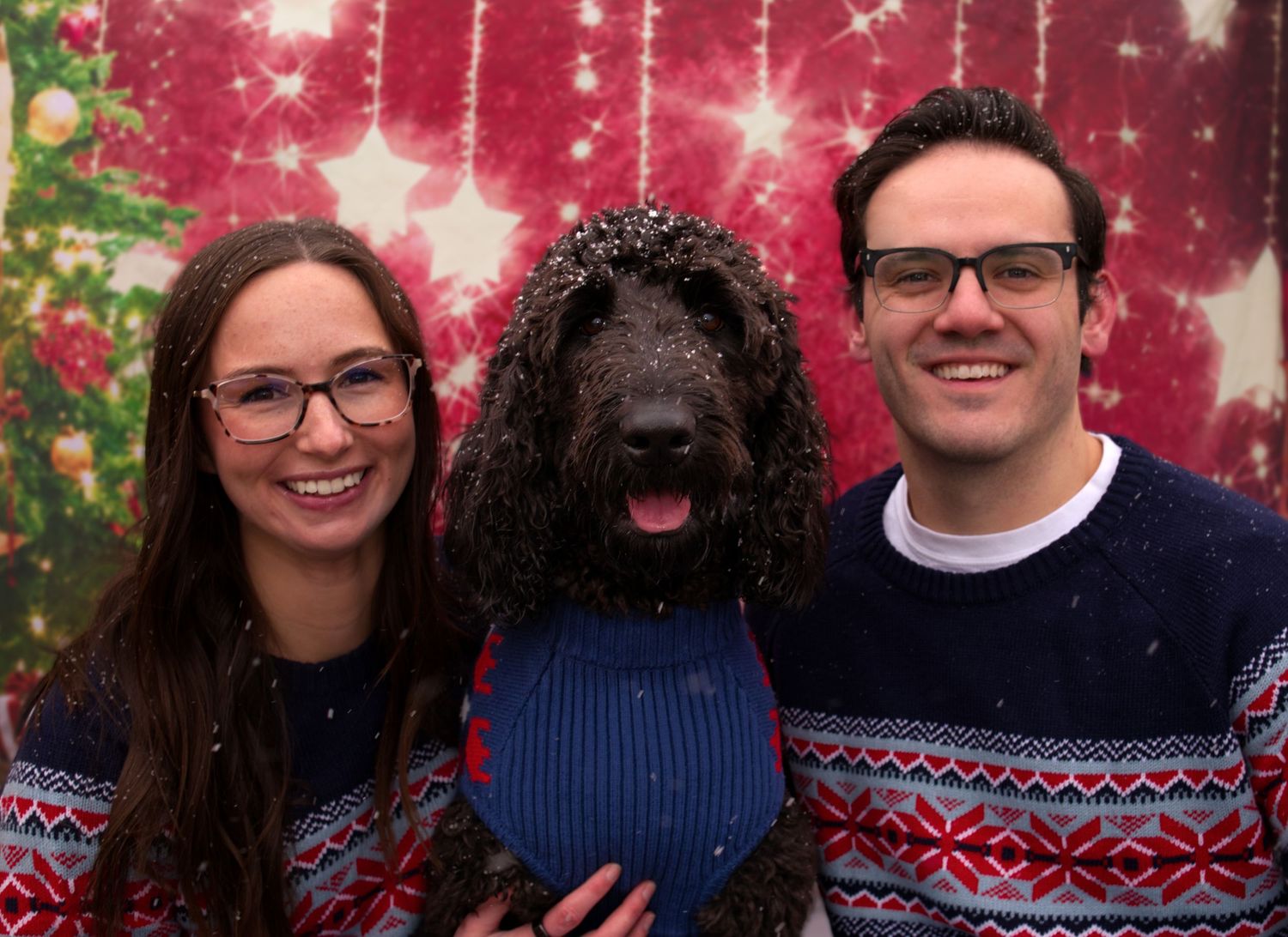 Couple in matching Christmas sweaters posing with their black curly-haired dog in front of festive holiday backdrop.