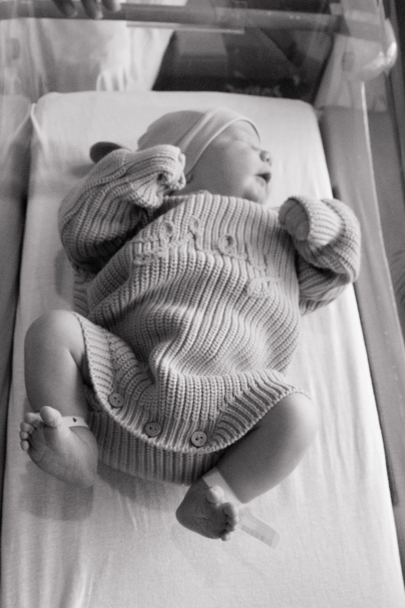 Black and white photo of a newborn baby in knit outfit lying in hospital bassinet.