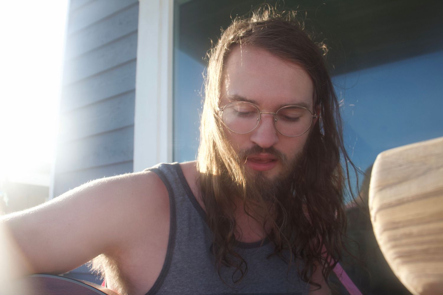 Long-haired bearded man with glasses playing acoustic guitar outdoors in warm sunlight.