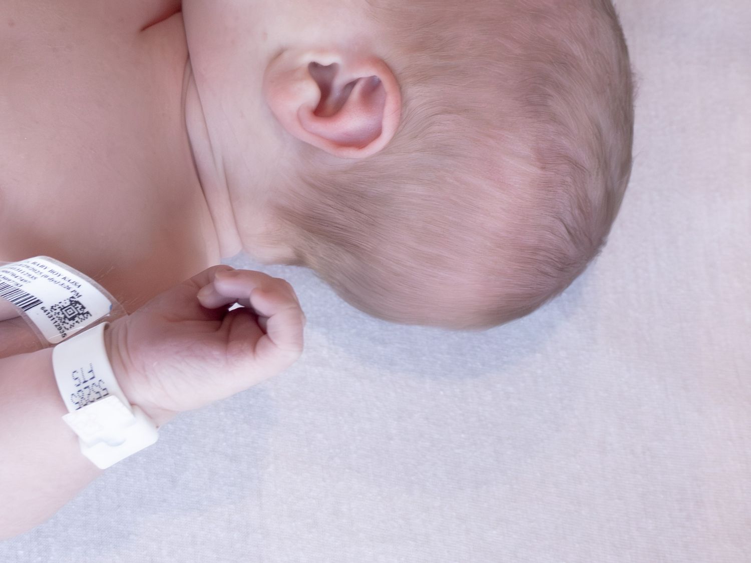 Newborn baby with hospital identification wristband lying on white blanket in maternity ward.