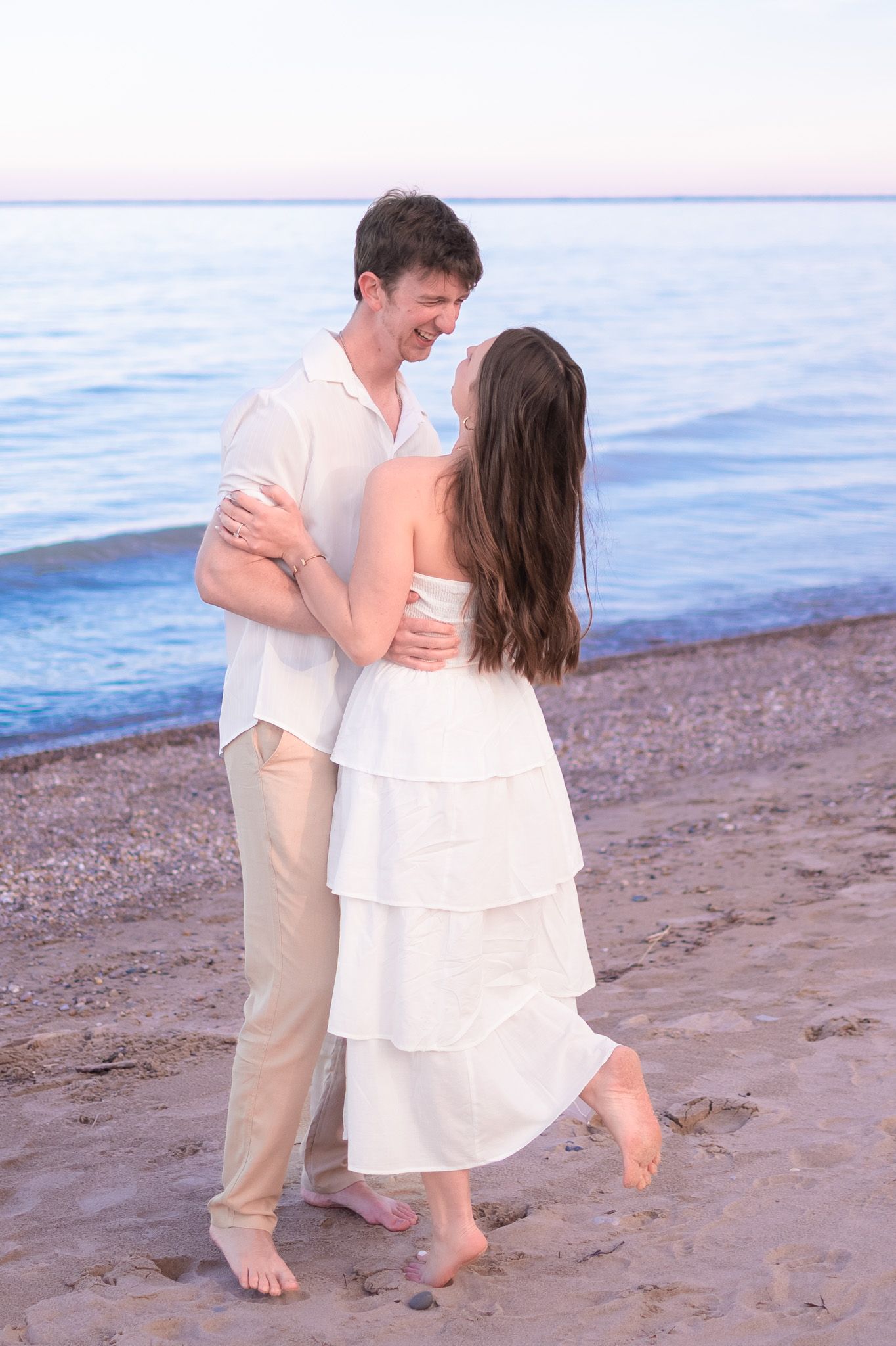 A romantic couple in white outfits embracing and smiling at each other on a peaceful beach at dusk.