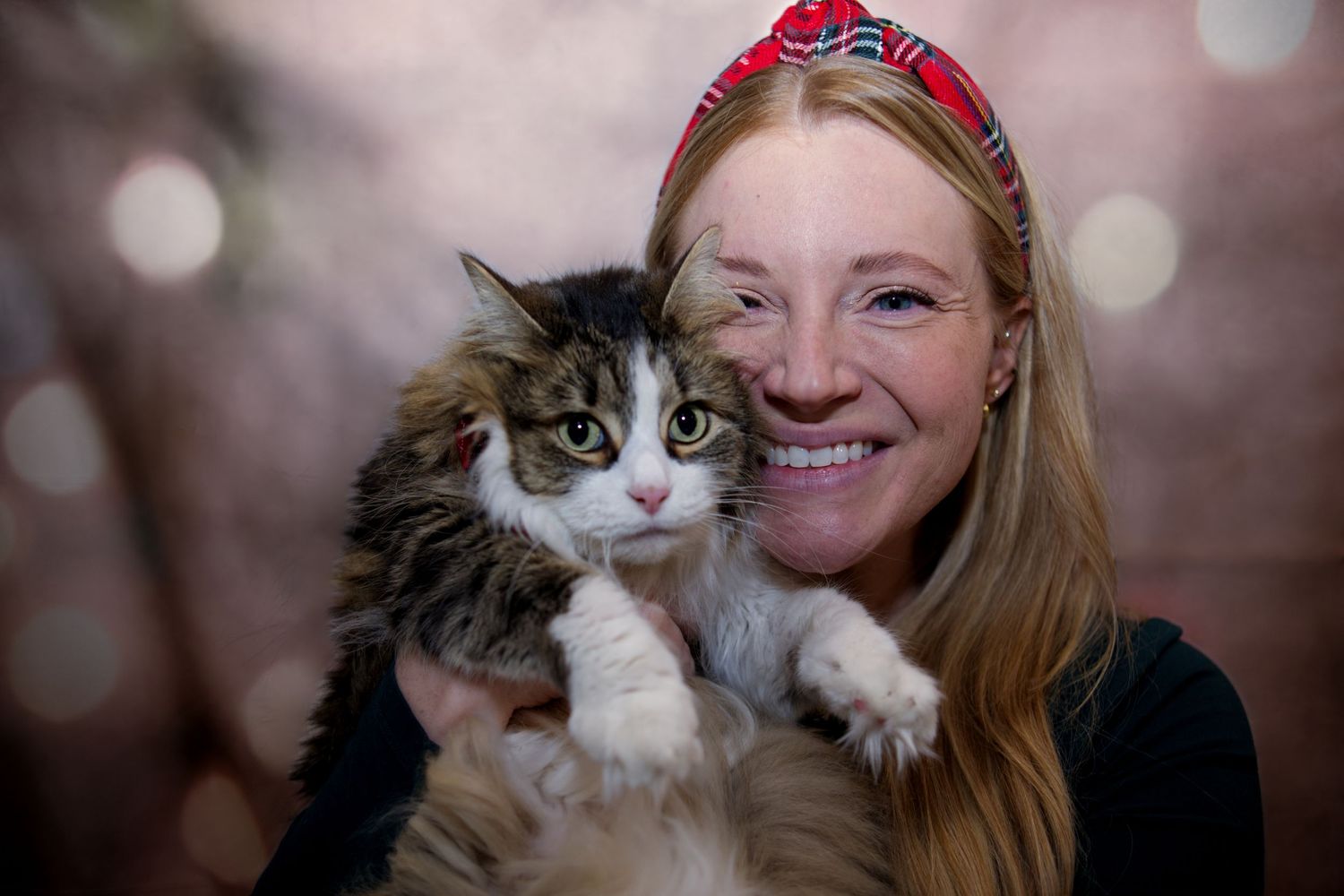 Smiling woman with red headband holding a fluffy tabby cat against a bokeh background.