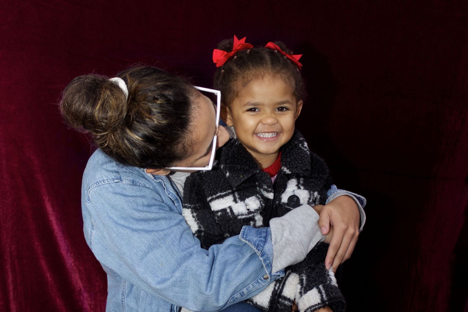 Woman in denim jacket hugging a smiling toddler girl with red bows in her hair.