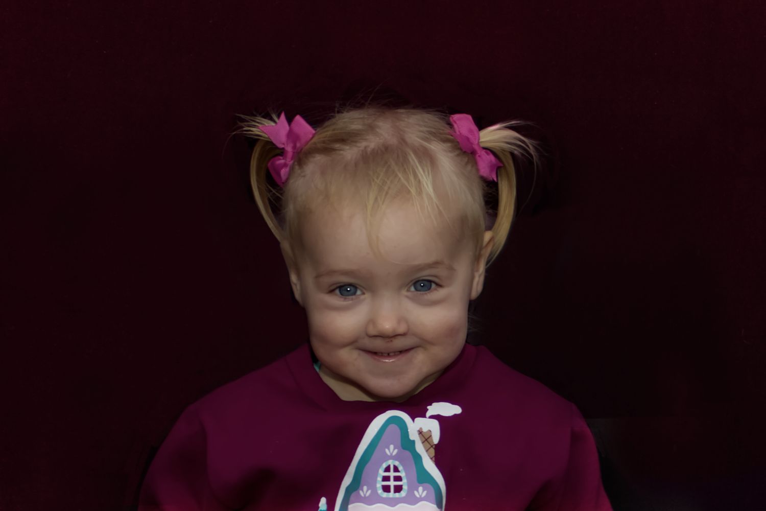 Adorable blonde toddler girl with pink pigtails smiling in a purple outfit against dark background.