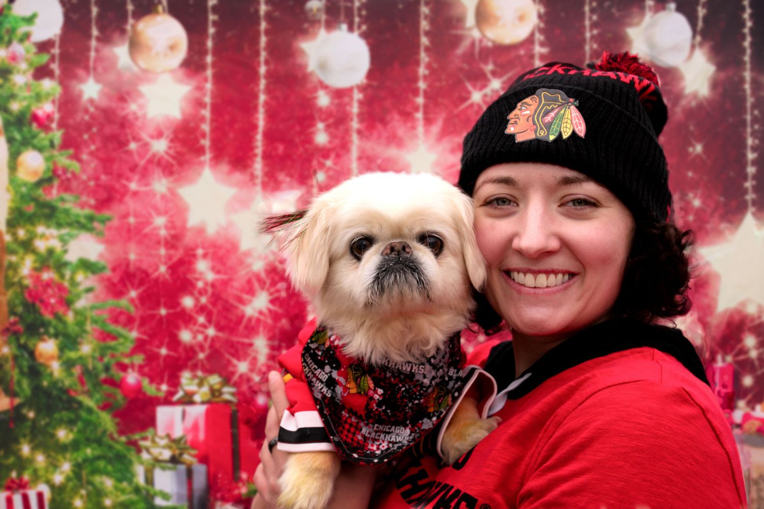 Smiling woman in Chicago Blackhawks hat holding small Pekingese dog dressed in holiday outfit at Christmas event.