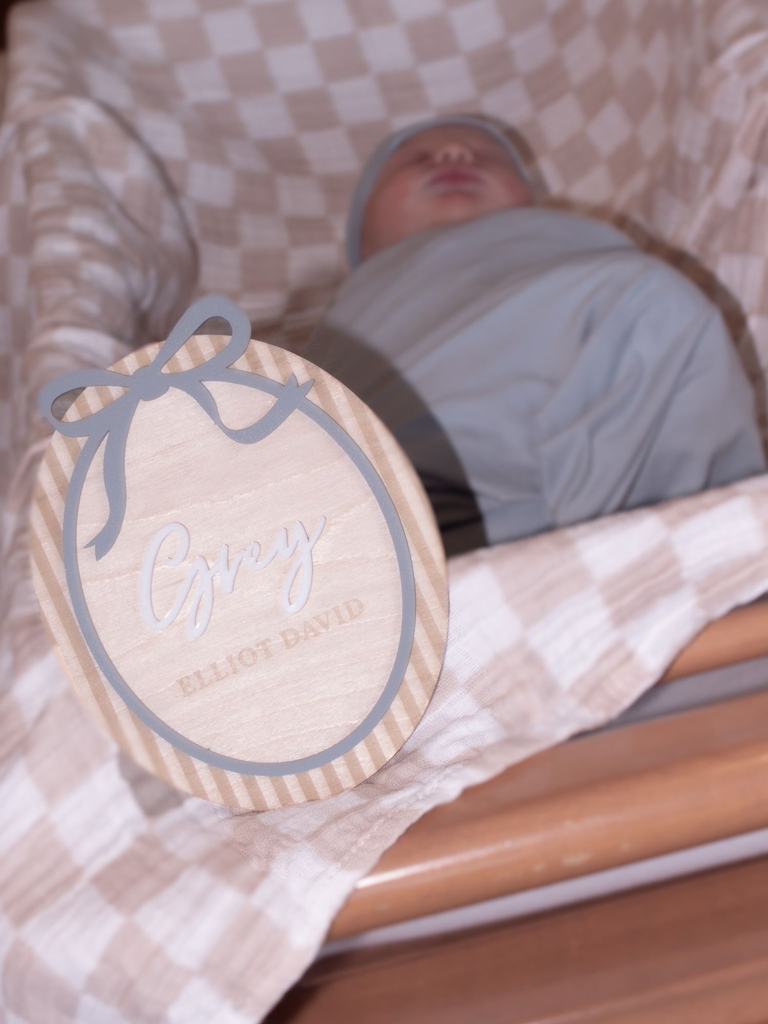 Sleeping newborn baby boy named Grey Elliot David in checkered blanket with name announcement sign.