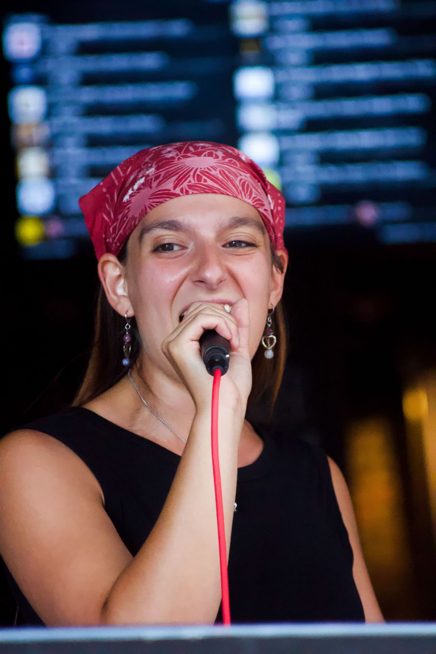 A young woman wearing a red bandana sings passionately into a red microphone at an indoor venue.