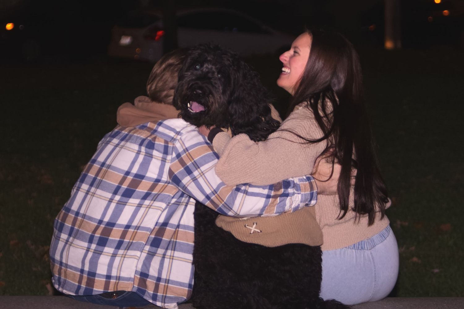 A couple sitting outdoors at night with their backs turned, embracing a black fluffy dog.