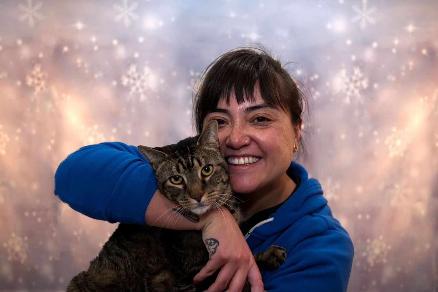 A smiling woman in a blue hoodie hugging a tabby cat against a snowy bokeh background.