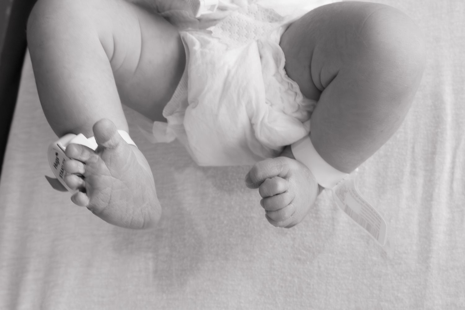 Black and white close-up of newborn baby feet and legs with hospital ID tag in maternity ward.