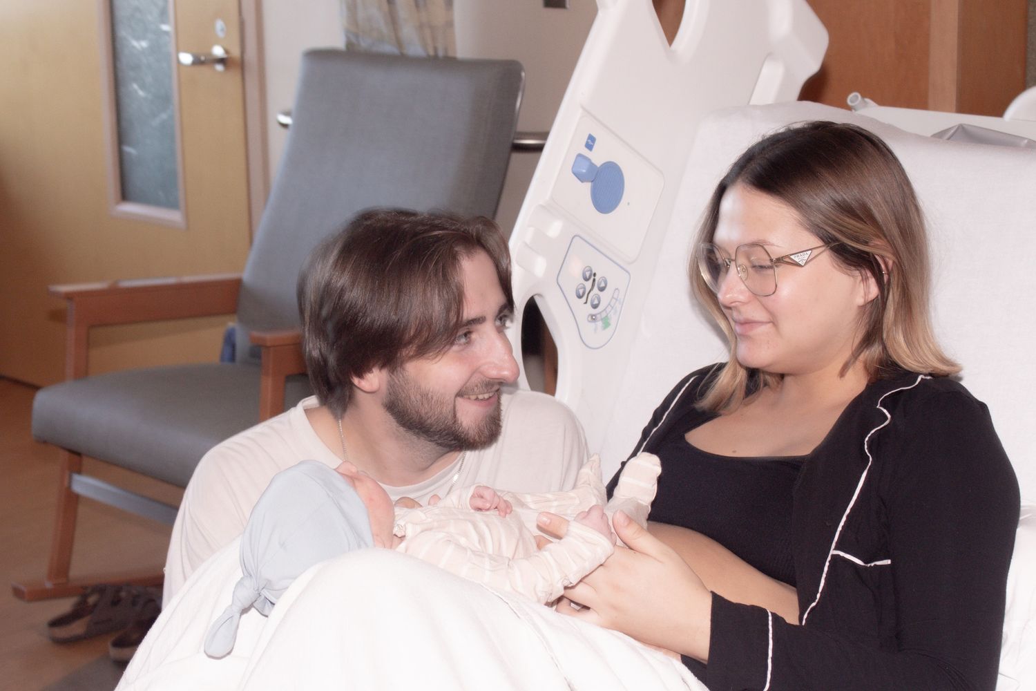 Happy new parents in hospital room smiling at each other while holding their newborn.