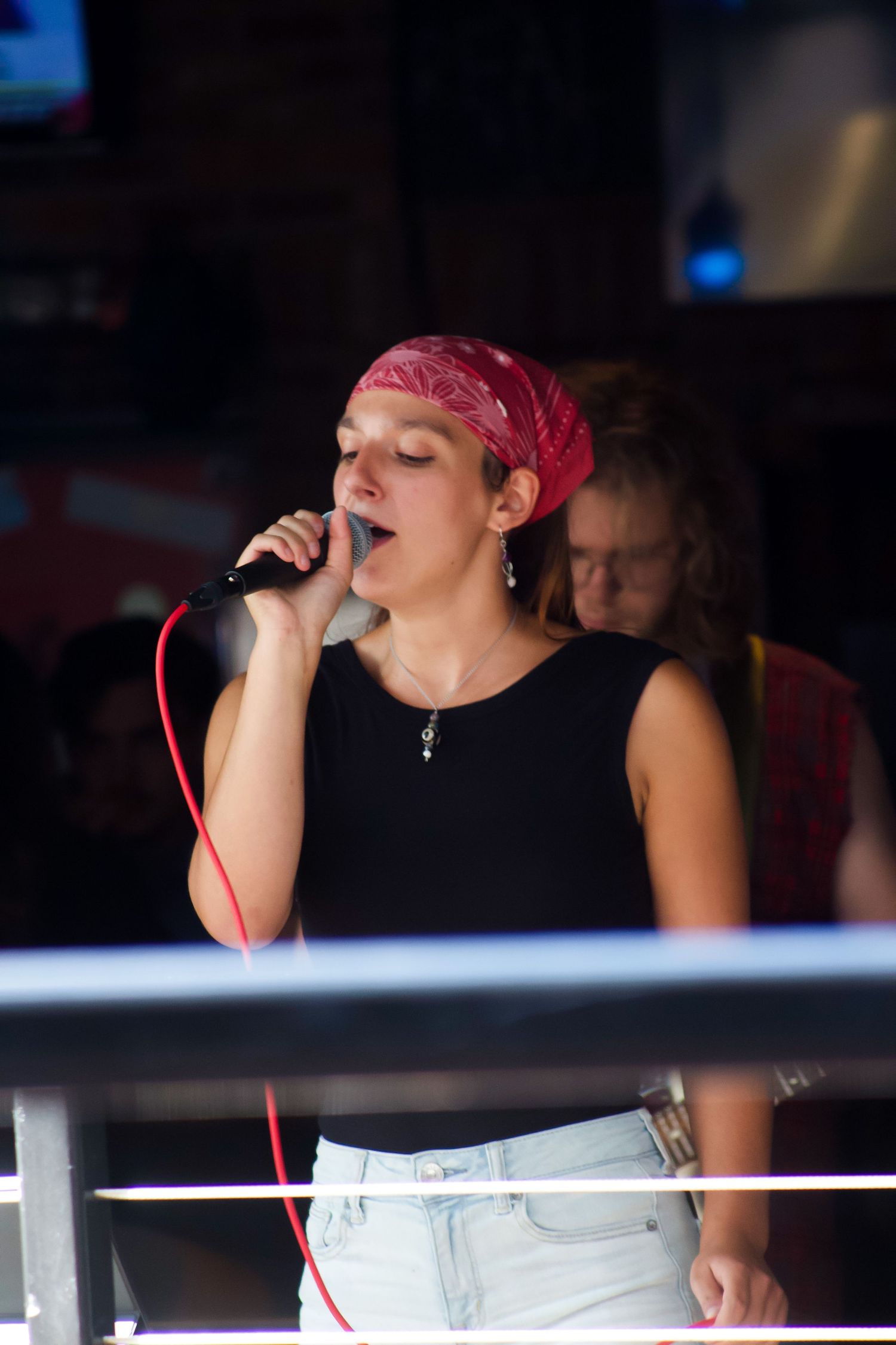 Young female vocalist singing into microphone wearing red bandana at indoor music venue.