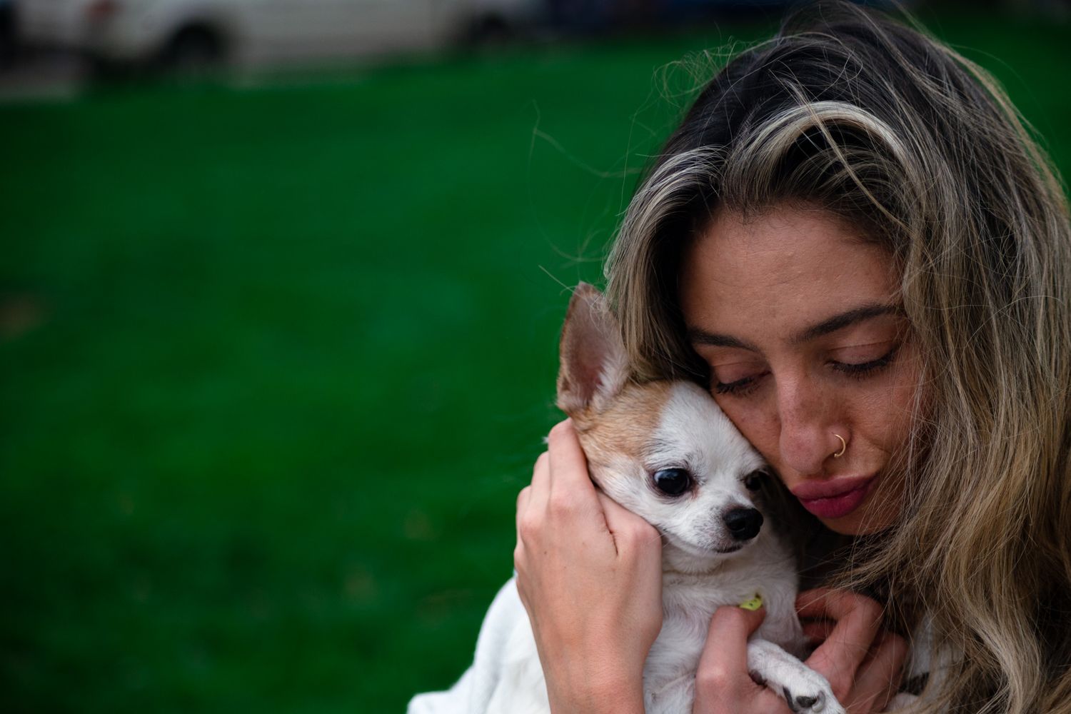 Woman with blonde highlights tenderly cuddling a small Chihuahua puppy against her cheek outdoors.
