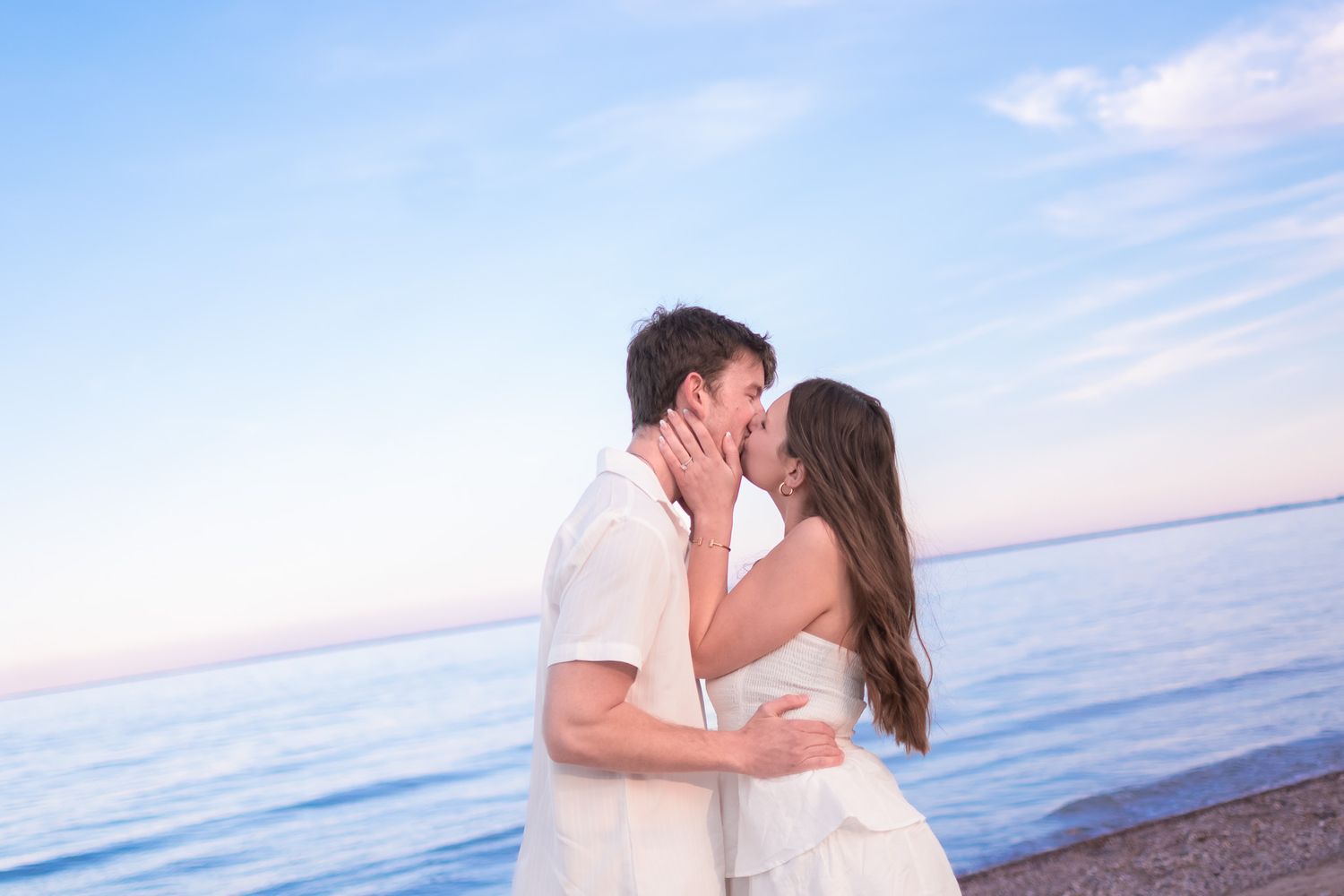 Couple sharing a romantic kiss on a beach at sunset with calm blue ocean behind them.