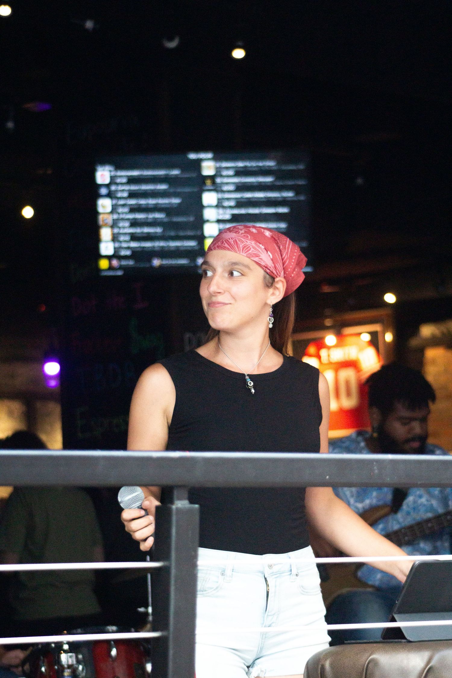 A young woman in a red bandana and black top holds a microphone while standing on an elevated stage.