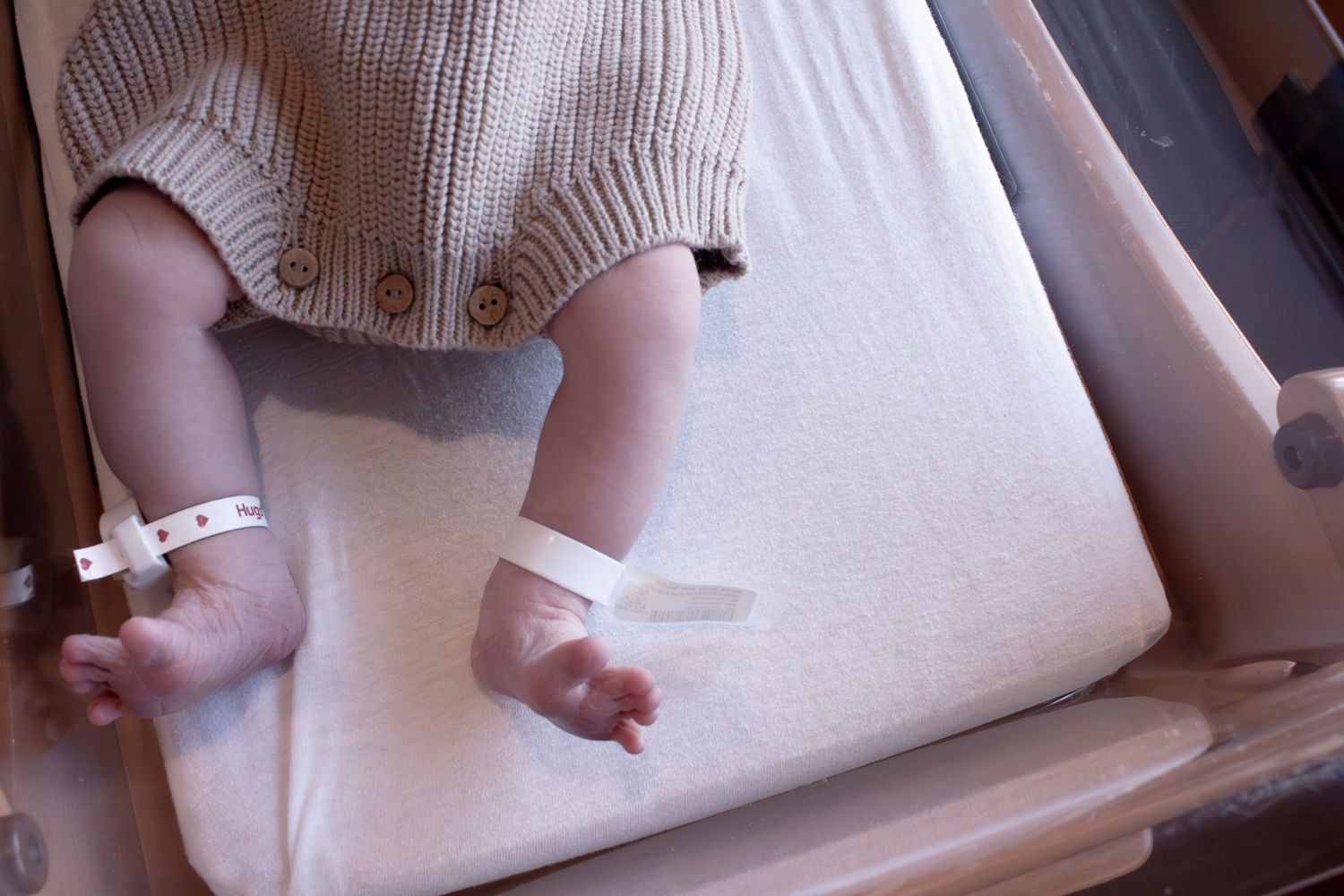 Newborn baby legs with hospital ID bracelets in knit onesie lying in bassinet.