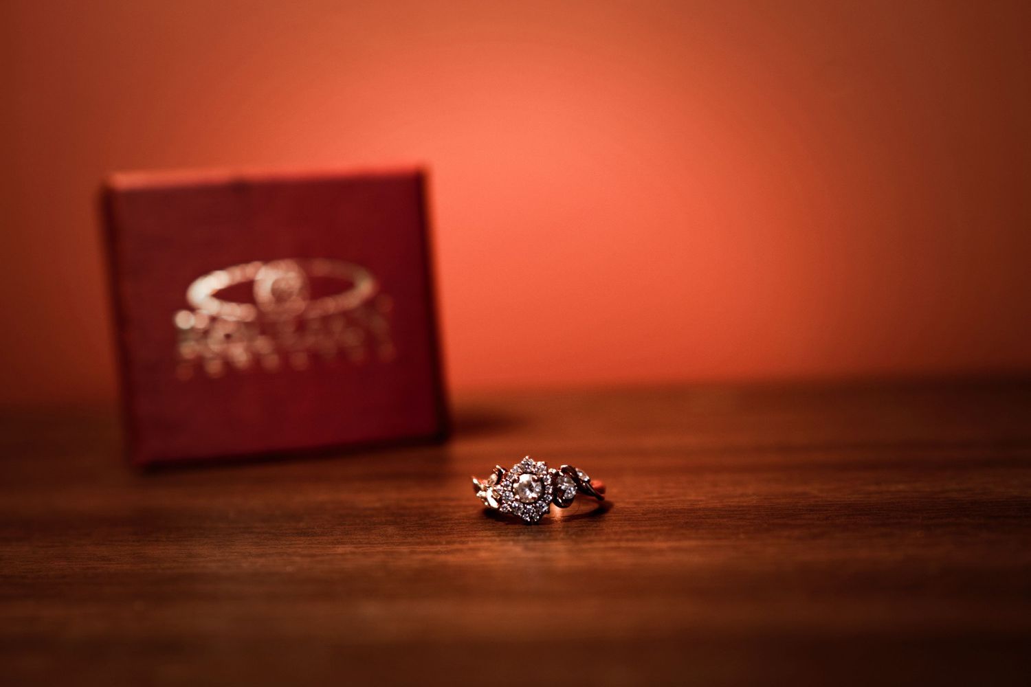 Diamond engagement ring beside a red jewelry box on a wooden table with warm orange background.