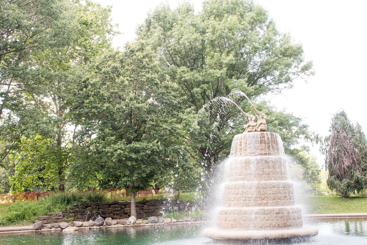 Ornate tiered stone fountain spraying water surrounded by lush green trees in a park setting.