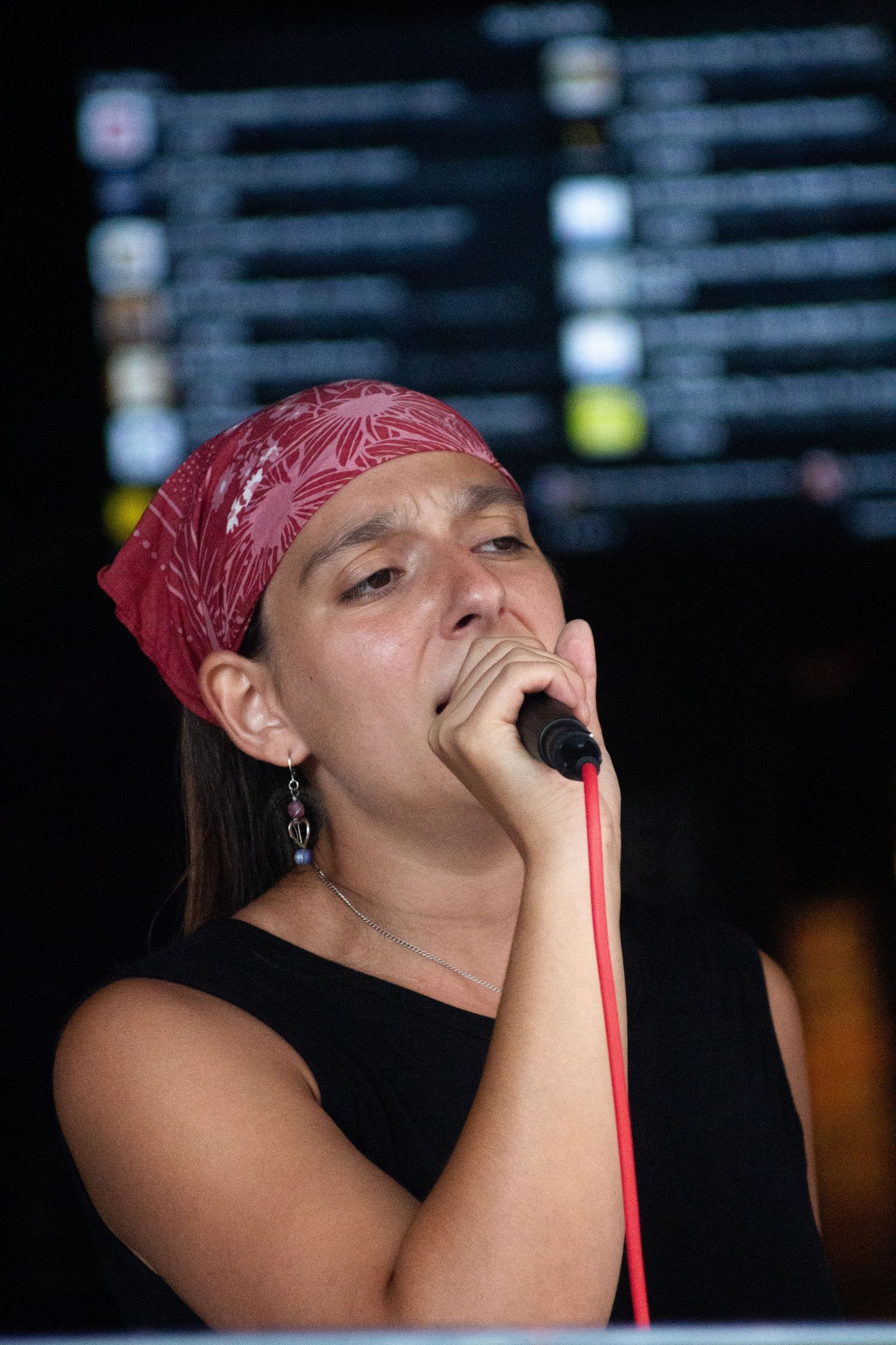A close-up of a female vocalist wearing a red bandana singing emotionally into a microphone at a live event.