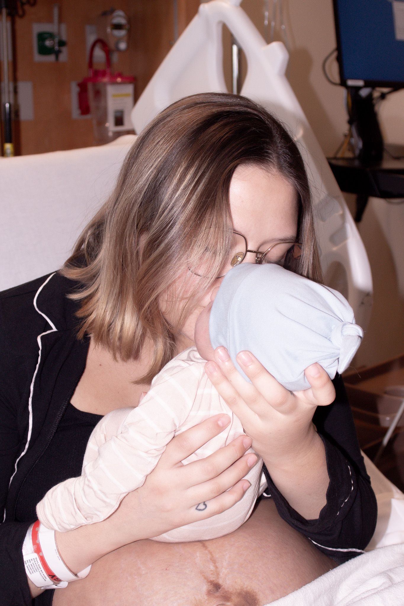 New mother in hospital bed kissing her swaddled newborn baby wearing a grey hat.
