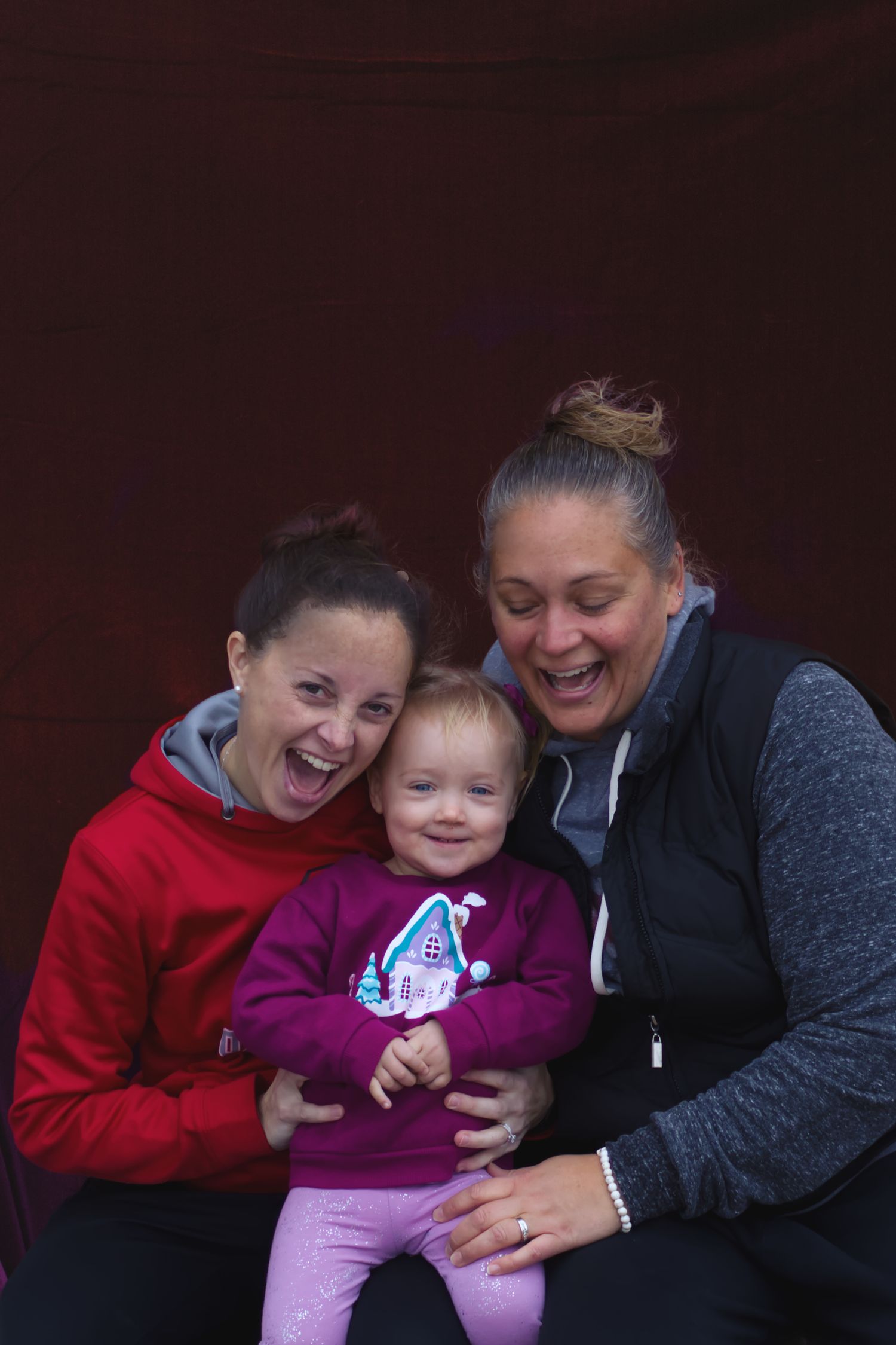 Two happy women laughing joyfully while holding a smiling toddler girl in a purple outfit.