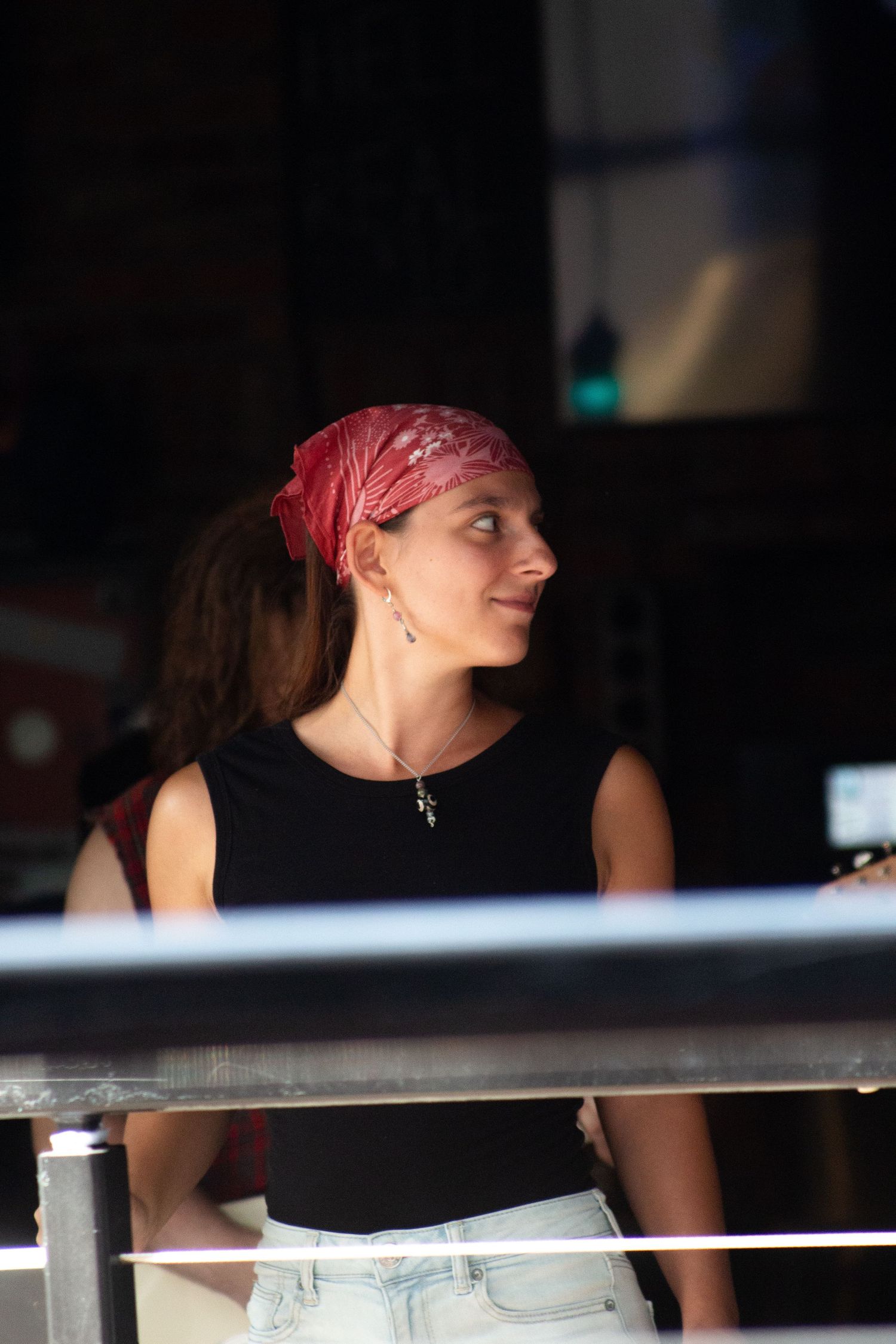Young woman in red bandana and black sleeveless top standing near railing at outdoor performance venue.