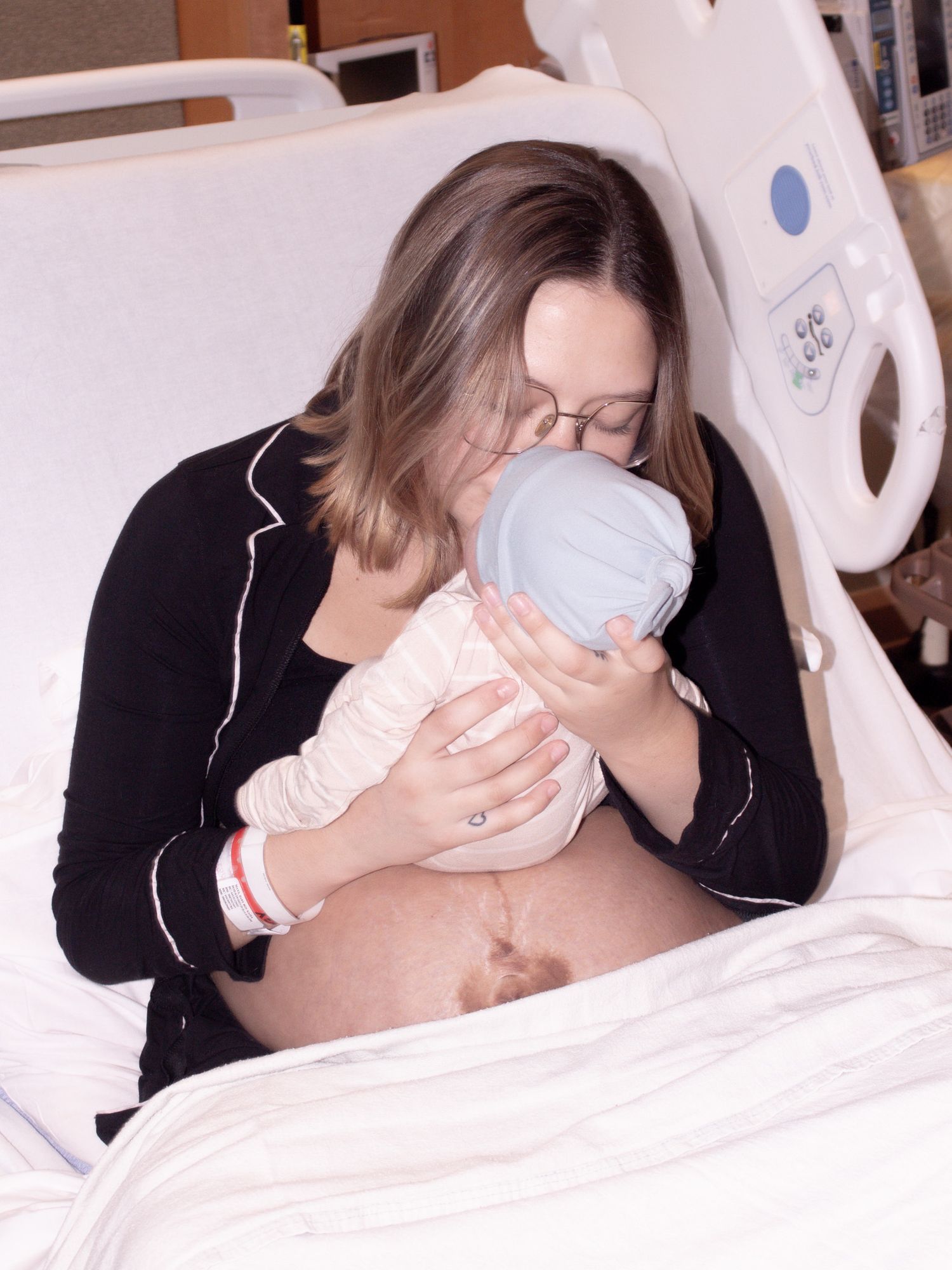 New mother in hospital bed kissing her newborn baby wrapped in a blue hat after delivery.