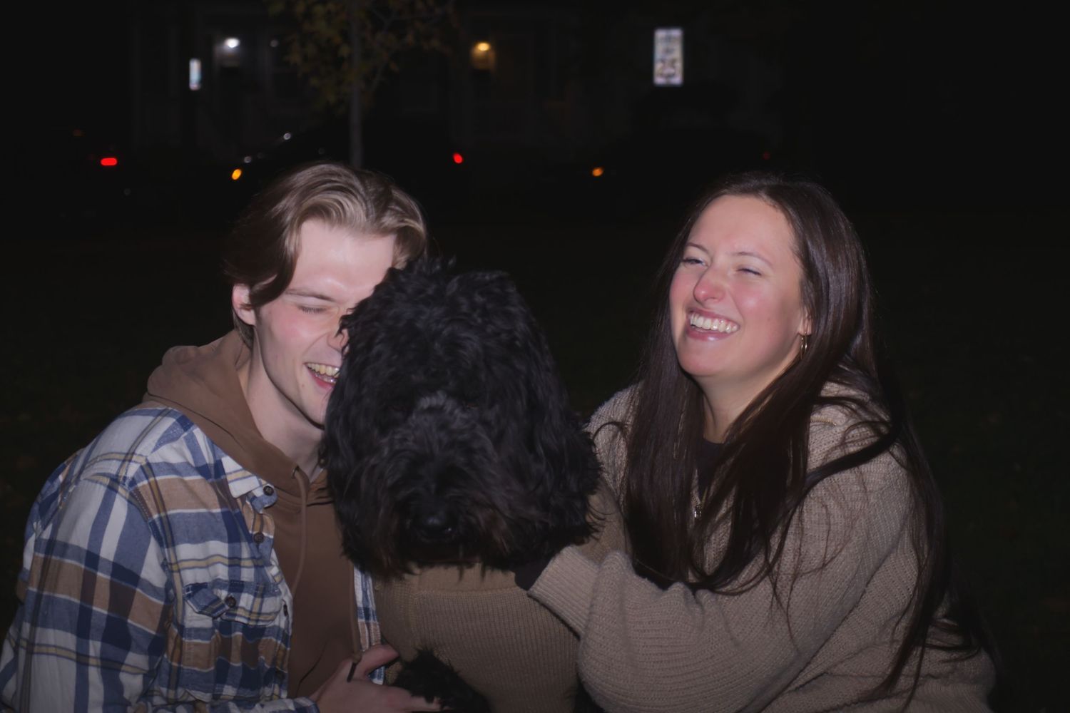 A young man and woman smiling while hugging a fluffy black dog outdoors at night.