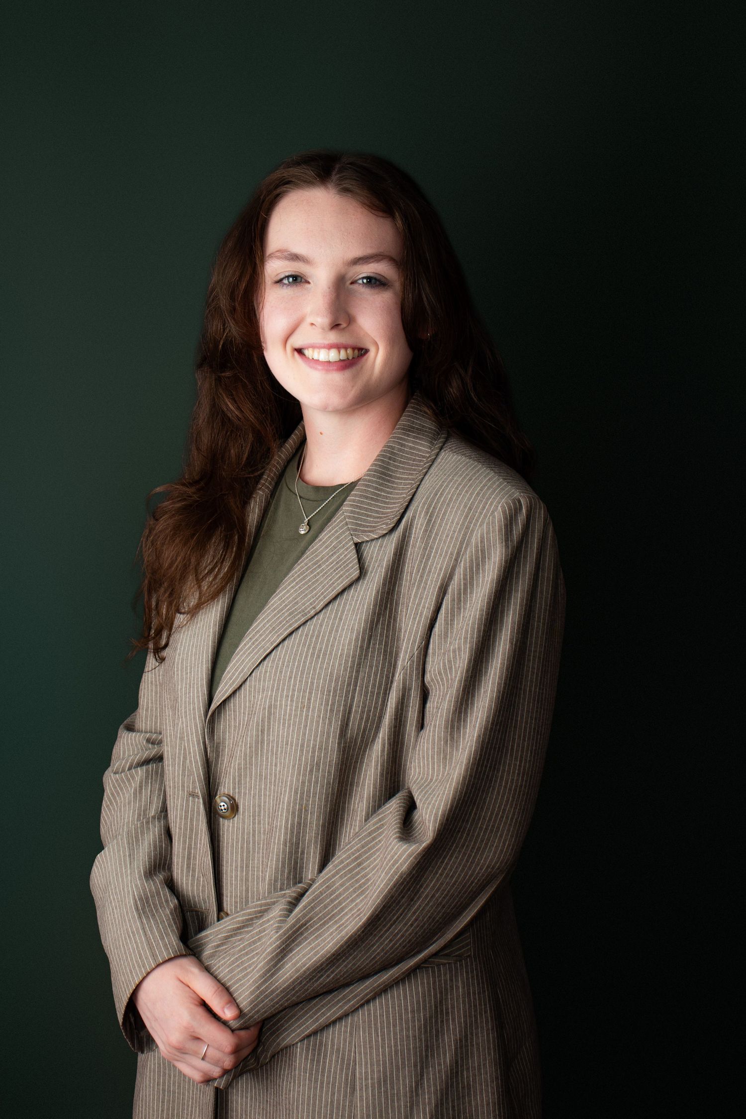 A smiling young woman in a striped blazer poses against a dark green background in a professional portrait.
