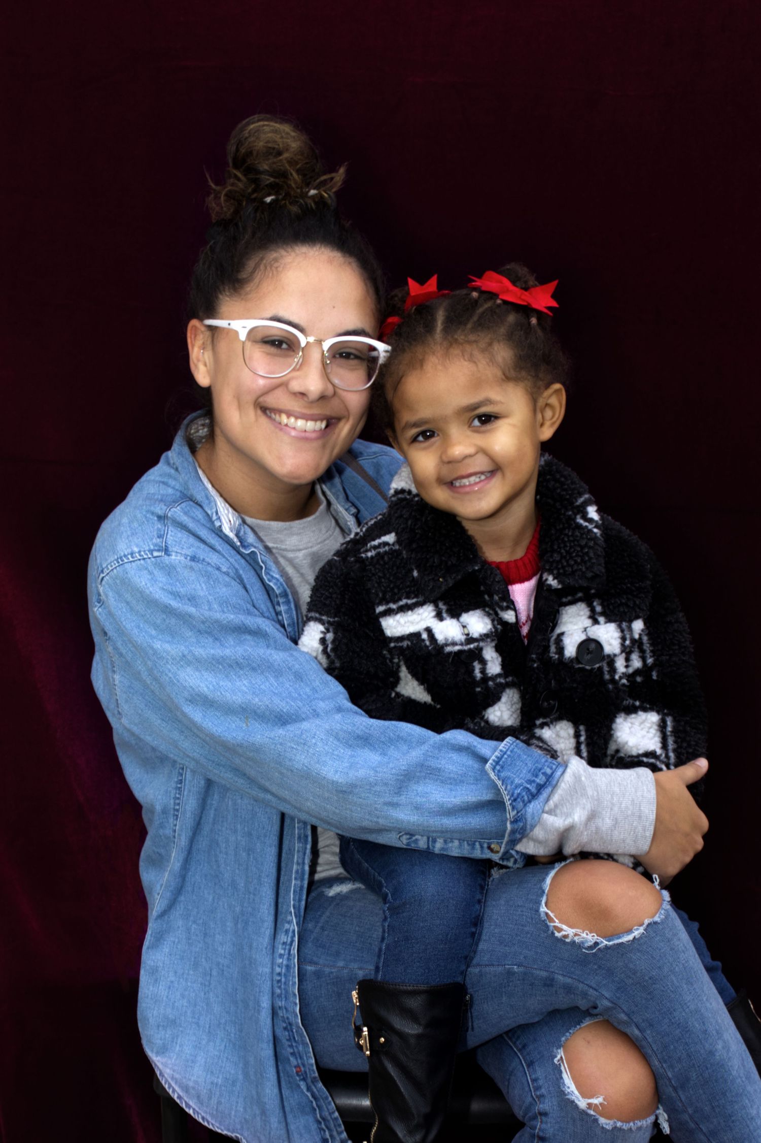 Smiling woman in denim jacket holding a young girl with red bow in her hair.