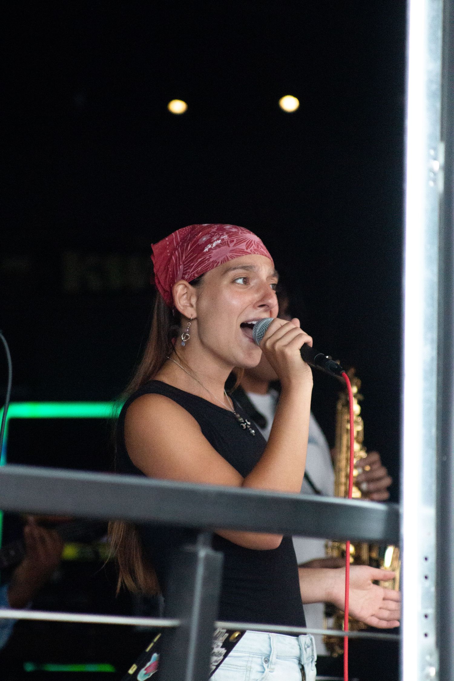 A female singer in a red bandana and black top performs on stage with a saxophone player visible behind her.