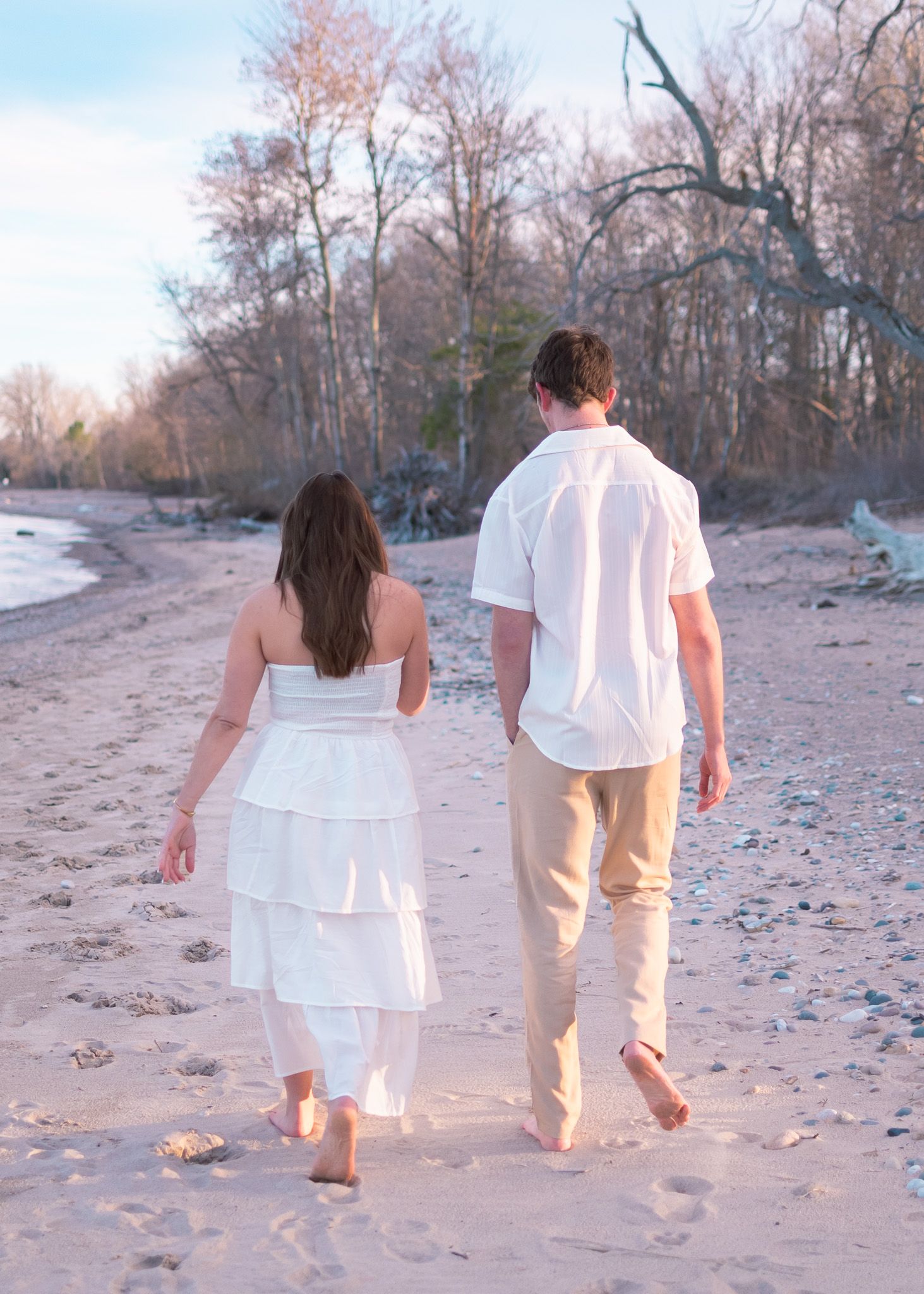 A couple in white outfits holding hands while walking barefoot along a sandy beach at sunset.