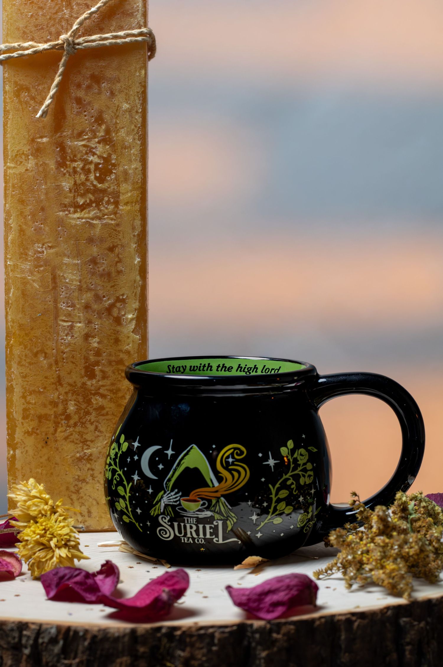 Black cauldron-shaped Suriel Tea Co. mug with green interior and floral design, beside dried flowers and a candle.