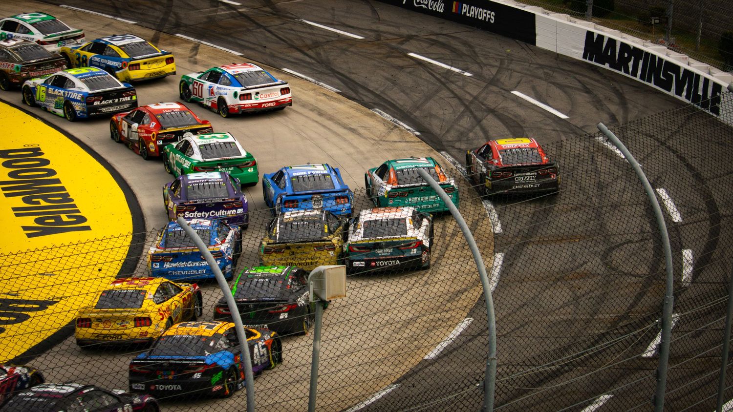 Aerial view of stock cars racing through a curved section of the speedway with yellow track markings.