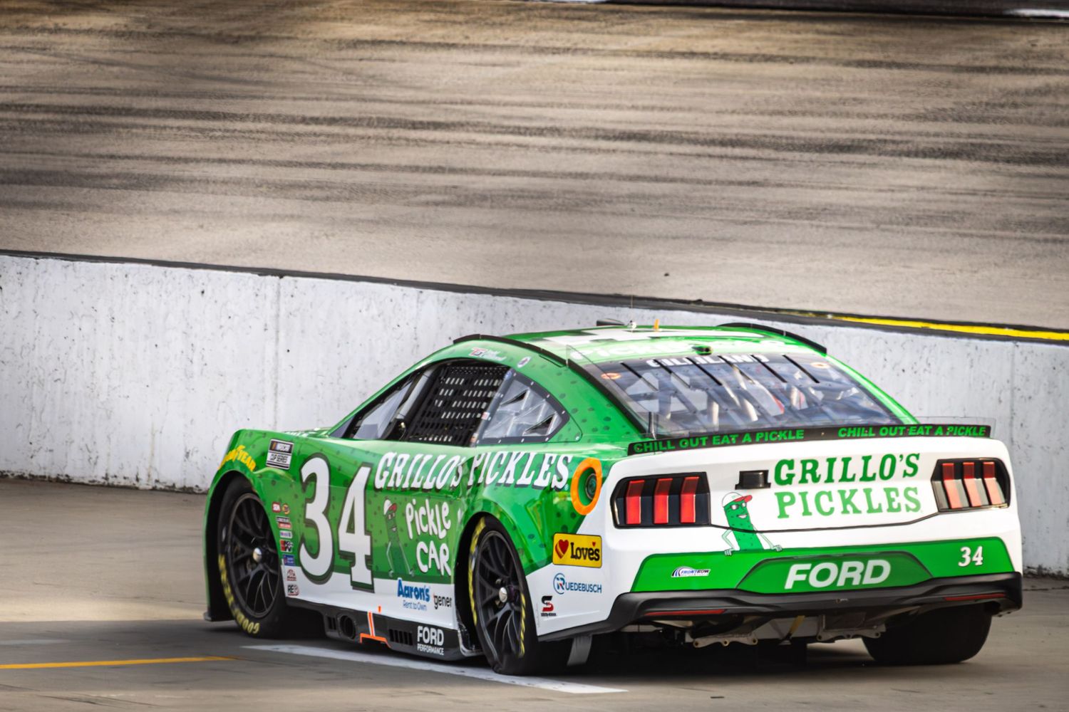 Row of NASCAR race cars speeding down a racetrack with green and white livery visible.