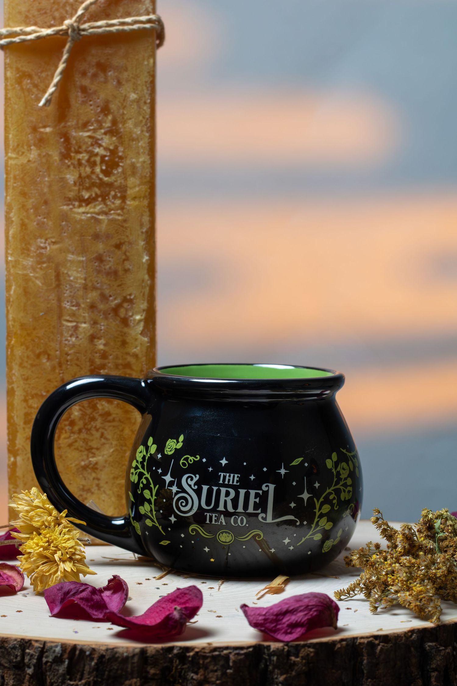 Black cauldron-shaped Suriel Tea Co. mug with green interior and floral design on rustic wood slice.