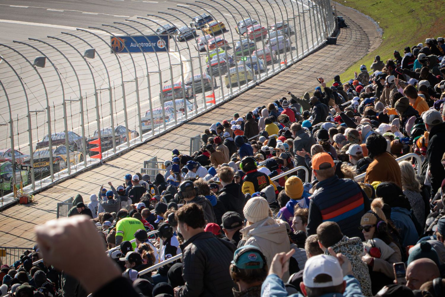 Curved racetrack with packed grandstands full of spectators watching motorsport action.