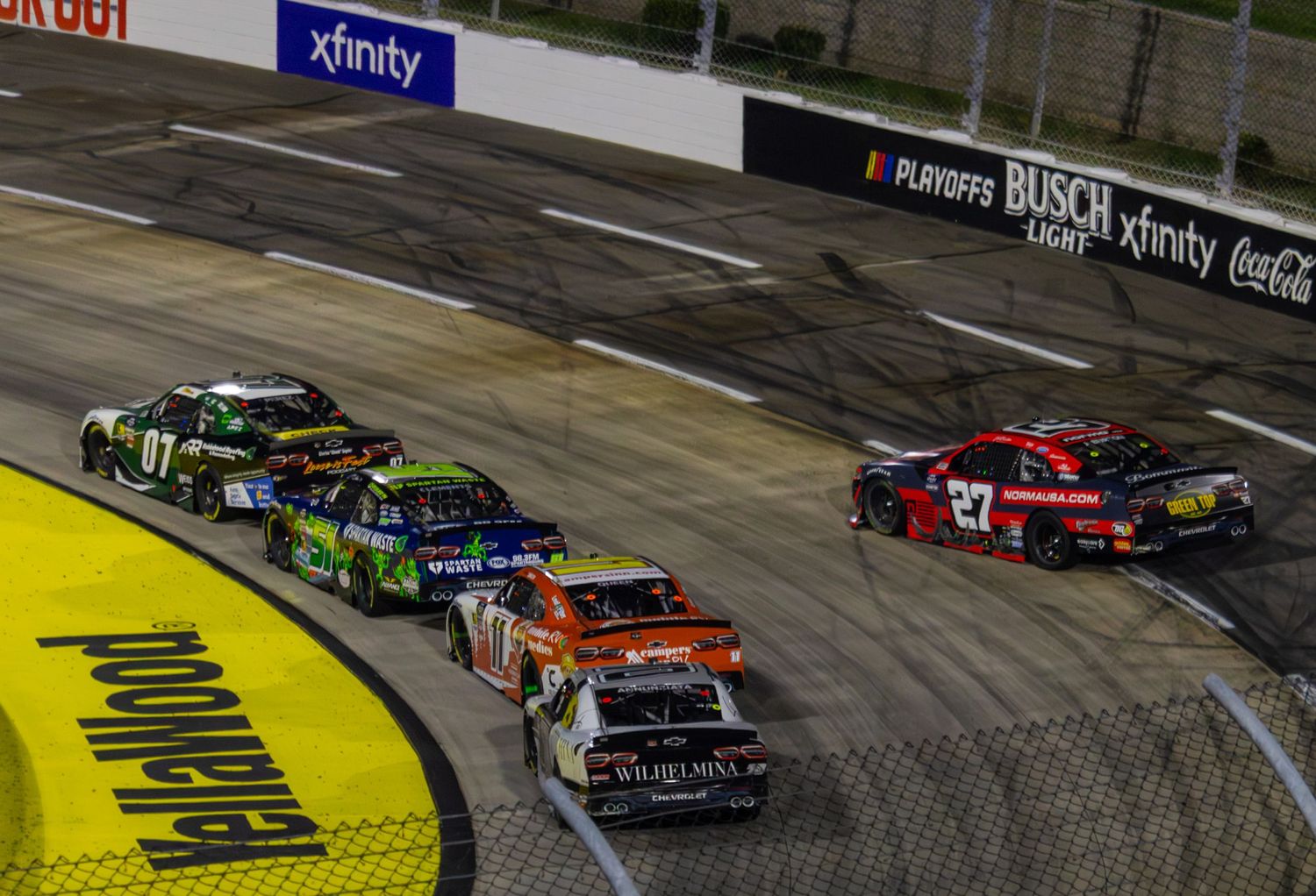 Pack of NASCAR vehicles race through turn with yellow curbing at Martinsville short track.