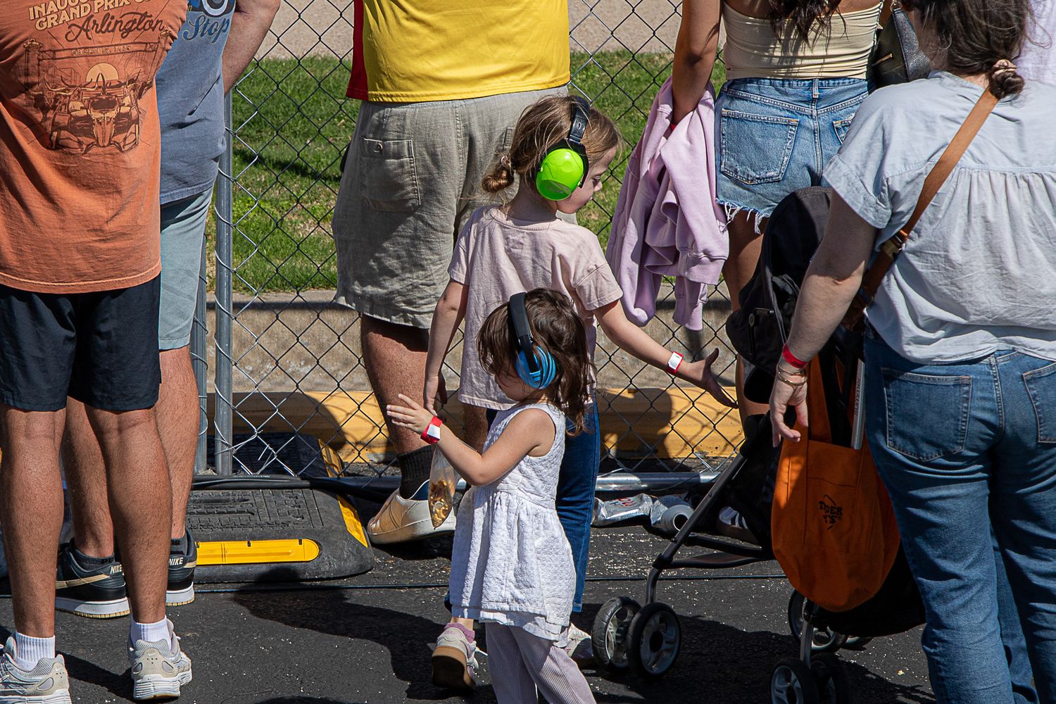 A crowd of people lined up along a chain link fence at an outdoor event on a sunny day.