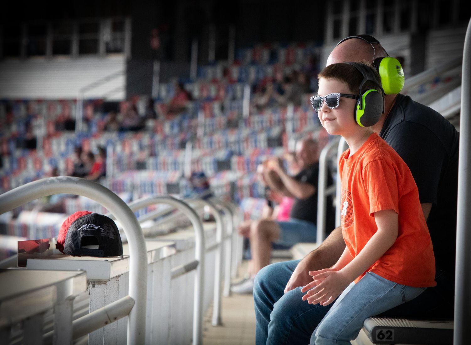 Spectators in bright safety gear watch intently from stadium seating at a motorsports event.